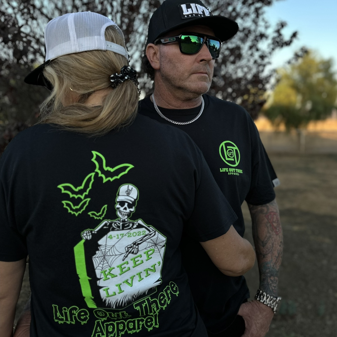 A man and woman stand outdoors, exuding a spooky vibe. The woman is wearing a cap and the "Unisex Keep Livin' T Shirt" by Life Out There Apparel, featuring a neon skeleton graphic and the text "Keep Livin'," making it perfect for Halloween. Beside her, the man sports sunglasses and a cap, along with a black shirt emblazoned with bold green letters. Trees and fields stretch out behind them.