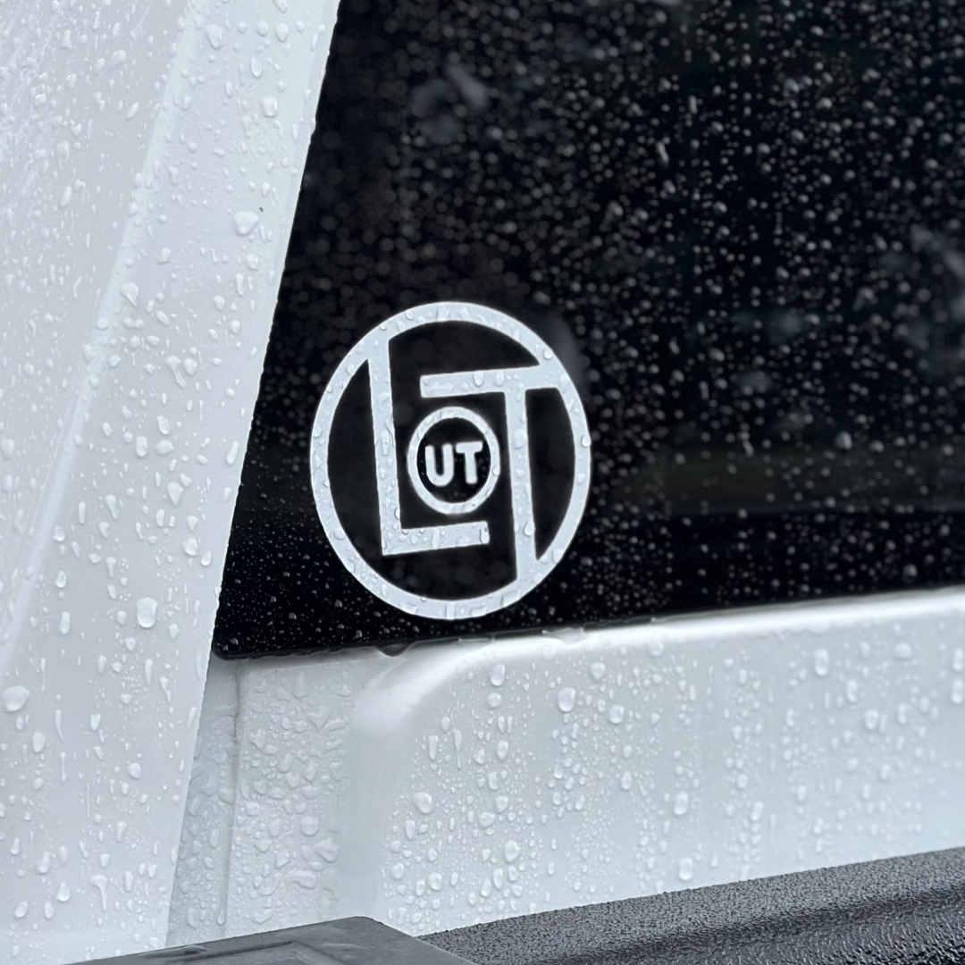 Close-up of a vehicle window with raindrops. A 3-inch die cut sticker from Life Out There Apparel is visible on the glass, showcasing the letters UT within a stylized design. The white vehicle's window frame is outlined in black.