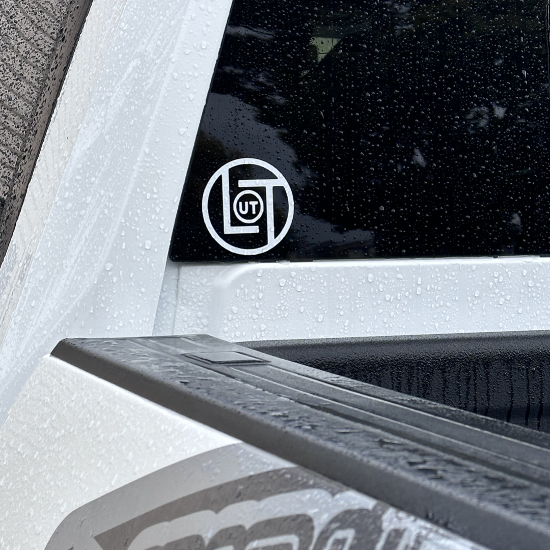Close-up of a vehicle displaying a 3-inch round die-cut sticker on the window, featuring an abstract design with a circle around "UT." The vehicle is wet with raindrops, and part of the truck's bed is visible. The sticker is from Life Out There Apparel, named LOT - Die Cut Sticker - 3 inch (White - Black - Red).