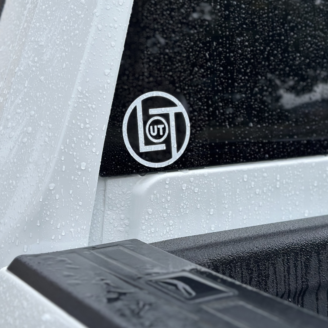 Close-up of a vehicle's rear window showcasing a 3-inch round die cut sticker from Life Out There Apparel, prominently displaying the letters UT at its center. The glass and surrounding surfaces are adorned with glistening water droplets, suggestive of a recent rain. The vehicle features a sleek white exterior accompanied by a textured black truck bed.