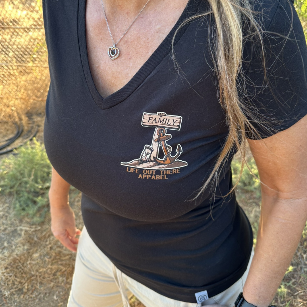 A person stylishly stands wearing the Time Flies Women's V-Neck T-Shirt in black from Life Out There Apparel, featuring a small logo with an anchor and rope, along with the words "FAMILY" and "LIFE OUT THERE APPAREL". They complete their look with a heart pendant necklace and light-colored pants. A backdrop of grass and fences suggests that time gently passes by in this peaceful setting.