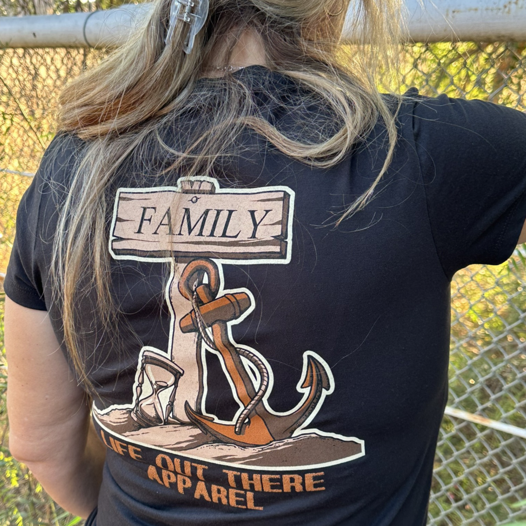 A person wearing a black V-neck T-shirt featuring an anchor design and the word "Family" stands in front of a chain-link fence. The shirt, named the Time Flies "Women's" V-Neck T Shirt by Life Out There Apparel, reflects the transient beauty of nature as trees and sunlight form a backdrop that serves as a reminder of life's fleeting moments.