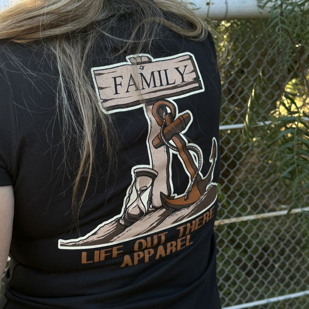 A person with long hair is wearing a black V-neck T-shirt adorned with an anchor, an hourglass, and a "Family" sign. The shirt is a Time Flies design by Life Out There Apparel. The phrase "TIME FLIES" hovers above the scene, which features a chain-link fence and lush foliage in the background.