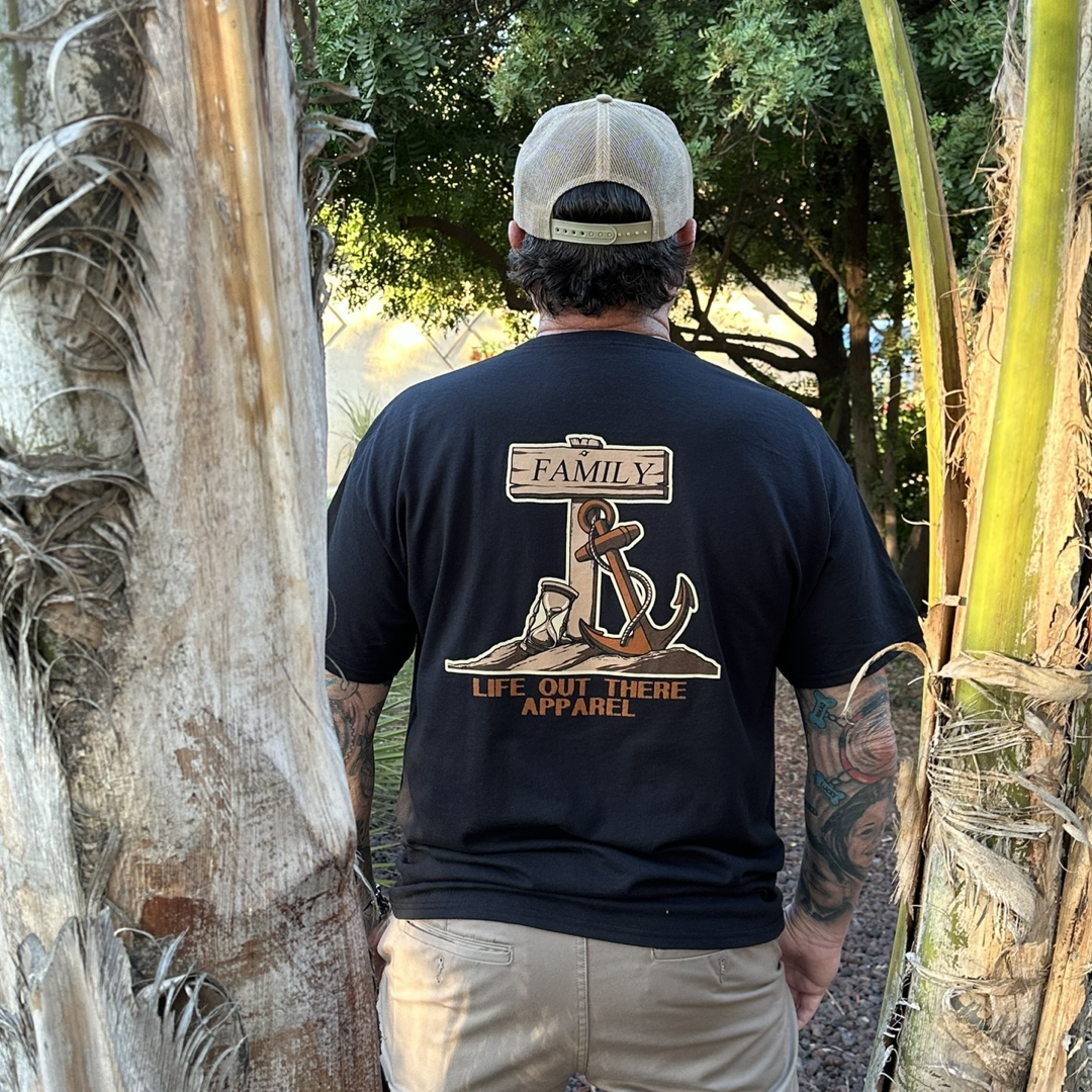 A person is seen from behind, sporting a black "Unisex Time Flies T Shirt" by Life Out There Apparel. The shirt showcases an anchor and rope, with the words "Time Flies" above. Below the design is the brand name "Life Out There Apparel," while palm trees gracefully frame the garden-themed illustration.