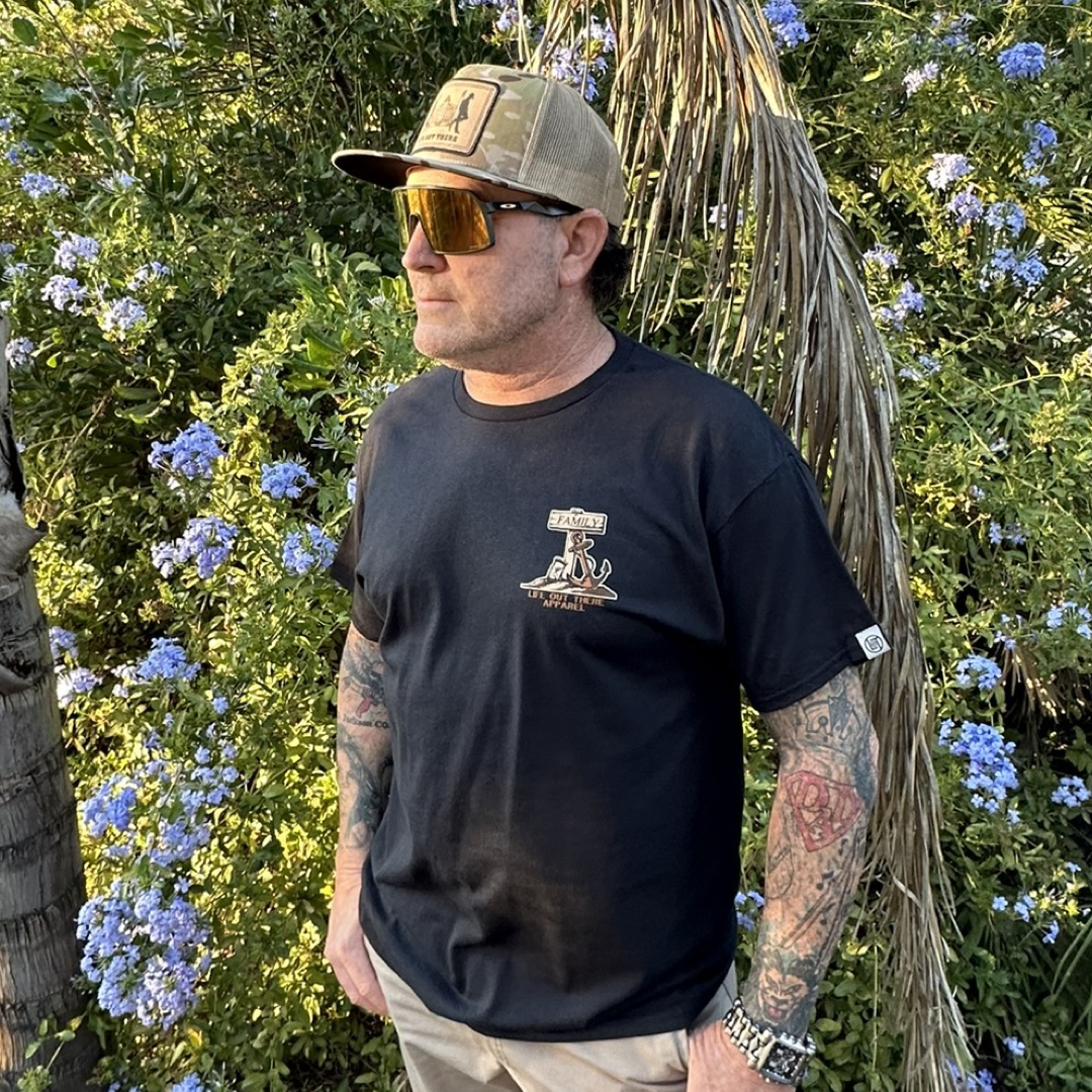 A man with tattoos, dressed in a Life Out There Apparel Unisex Time Flies T Shirt in black and sporting sunglasses, stands before a lush green backdrop adorned with blue flowers. He's donning a beige baseball cap and gazes to his left, seemingly contemplating how time slips away like sand through an hourglass.