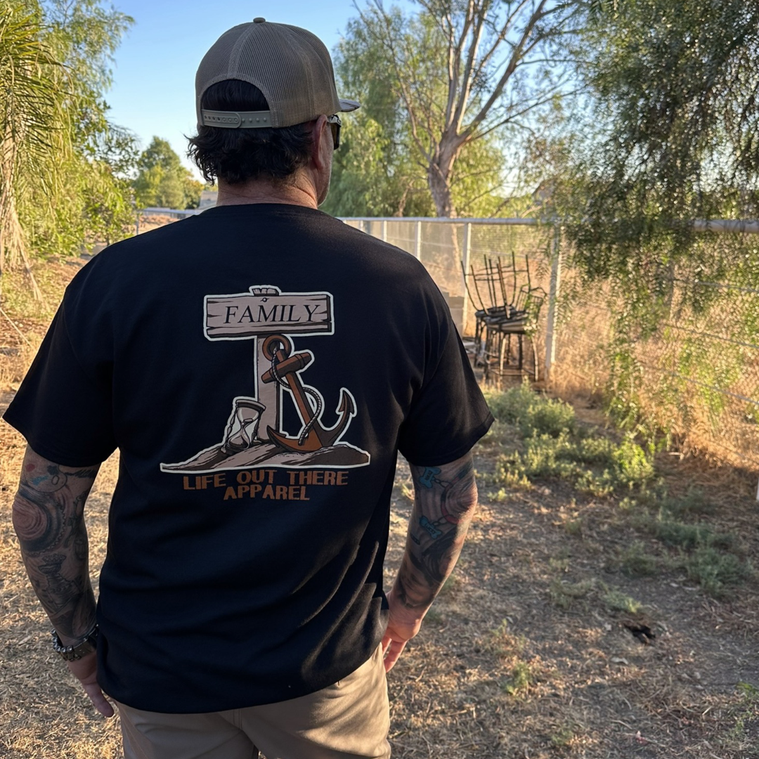 A man with tattoos stands outdoors facing away, wearing the Life Out There Apparel's Unisex Time Flies T-Shirt in black, which features an anchor design with "Family" above it. As time flies on this sunny day, trees and a pile of stacked chairs can be seen in the background.