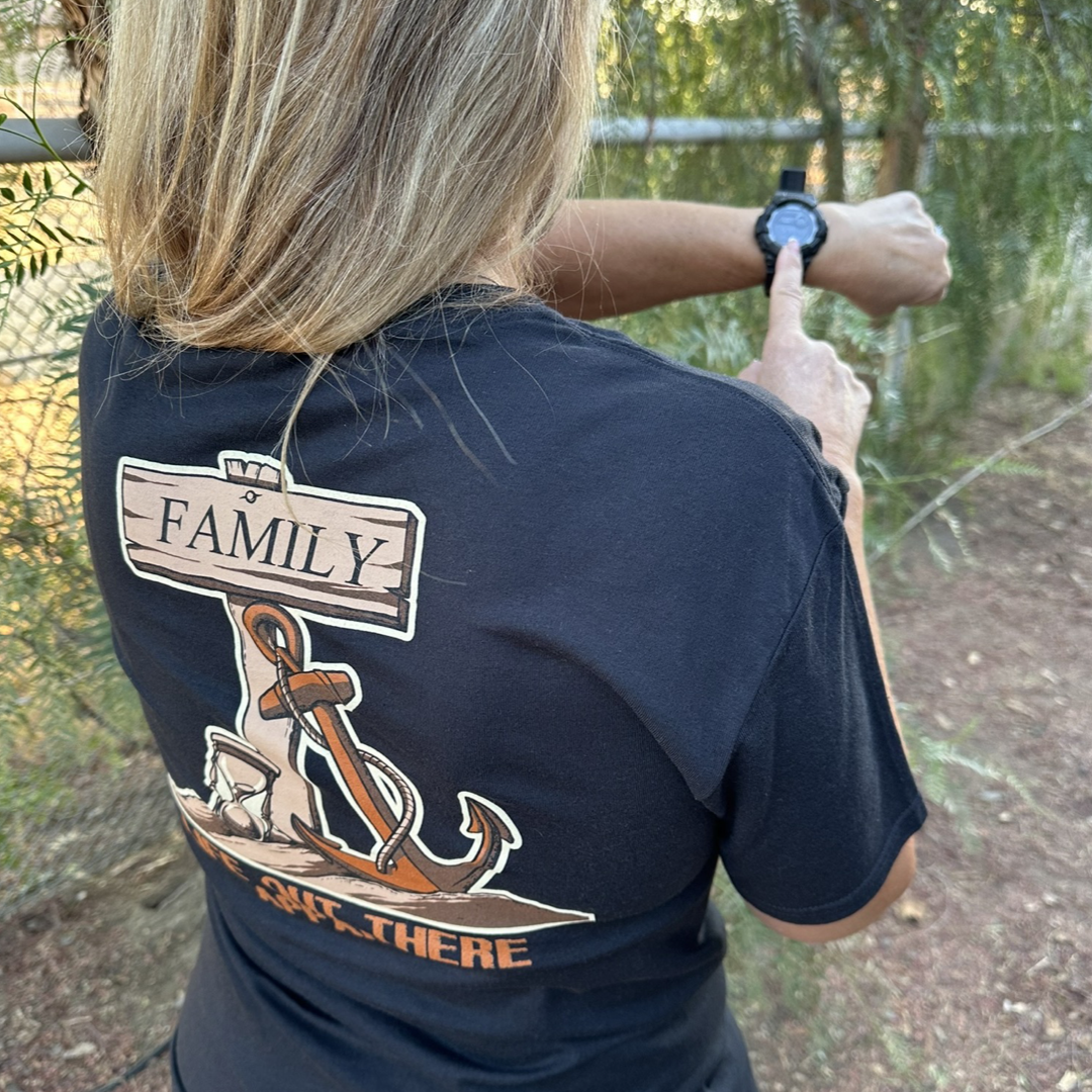 A person with long hair is seen outdoors wearing the Unisex Time Flies T Shirt in black from Life Out There Apparel, featuring an anchor design and the word "Family." They gesture towards a smartwatch on their wrist, subtly suggesting that TIME FLIES, all while facing away from the camera.