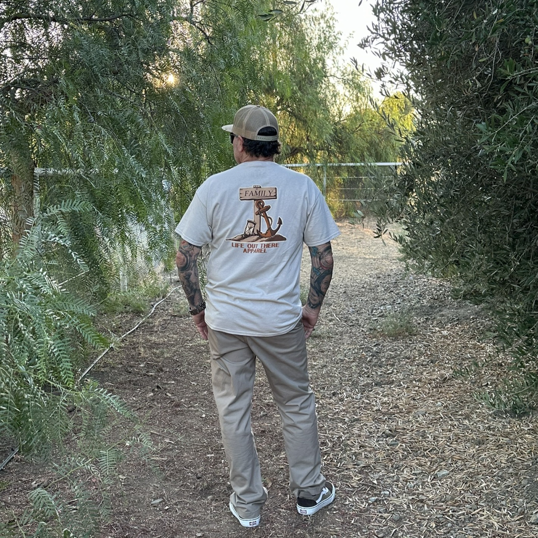 A man stands outdoors on a dirt path surrounded by trees, wearing a beige baseball cap, the Unisex Time Flies T-Shirt in Sandstone from Life Out There Apparel featuring an hourglass design on the back, and khaki pants. The setting sun casts a warm glow through the foliage, reminding us that time flies in such serene moments.