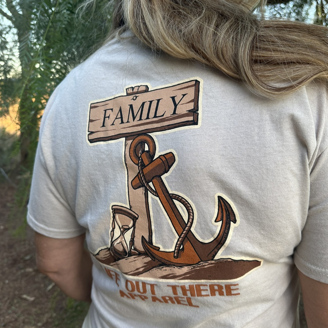 A person with long hair is wearing the Unisex Time Flies T-Shirt in sandstone, showcasing a sailor-themed design with an anchor, a "Family" sign, and various nautical elements. An hourglass subtly illustrates the passage of time. The bottom partially displays the words Life Out There Apparel.