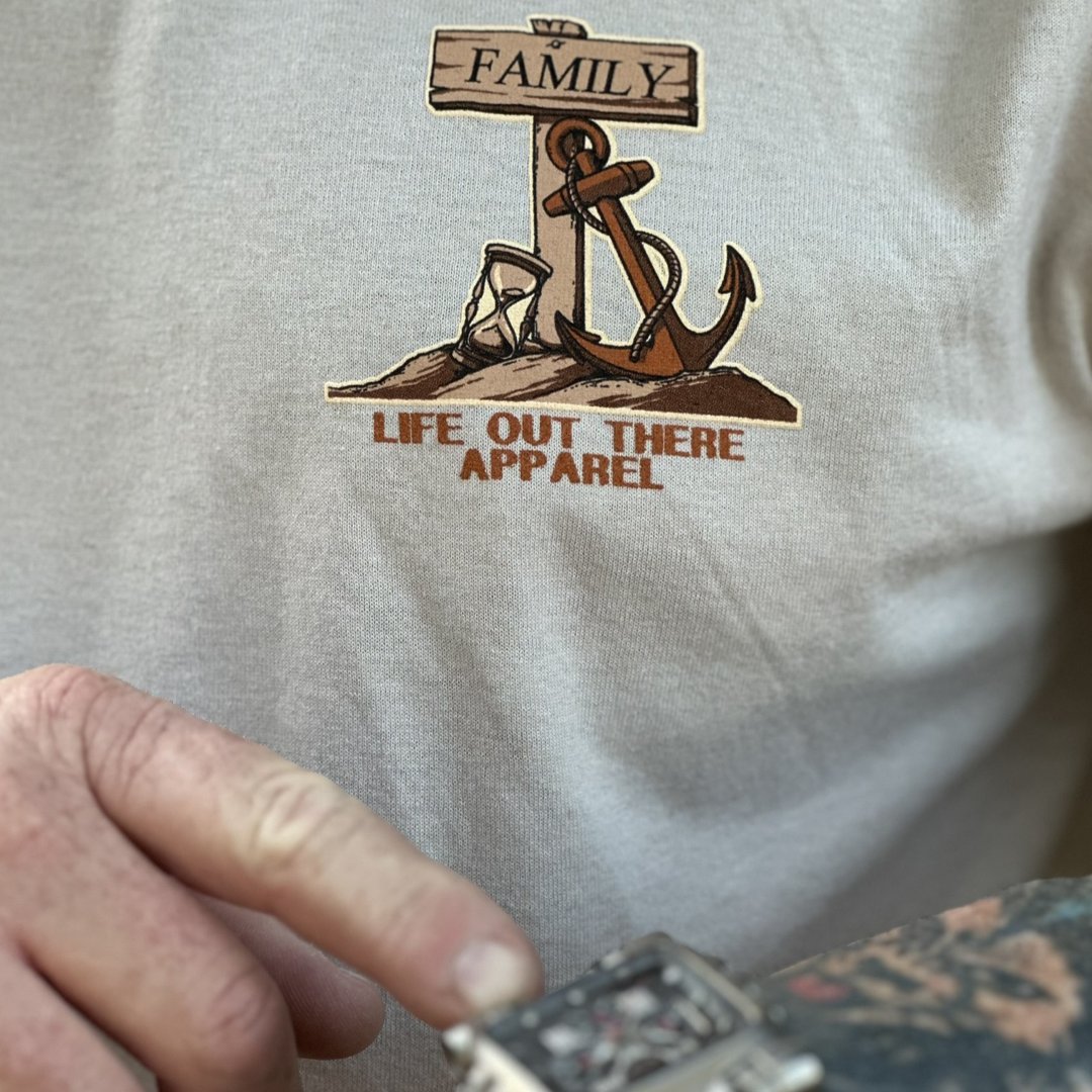 A person wearing the Unisex Time Flies T Shirt in sandstone, designed with an anchor, hourglass, and a wooden sign reading "Family," showcases the brand Life Out There Apparel. To highlight how time flies, they adjust a watch on their wrist.
