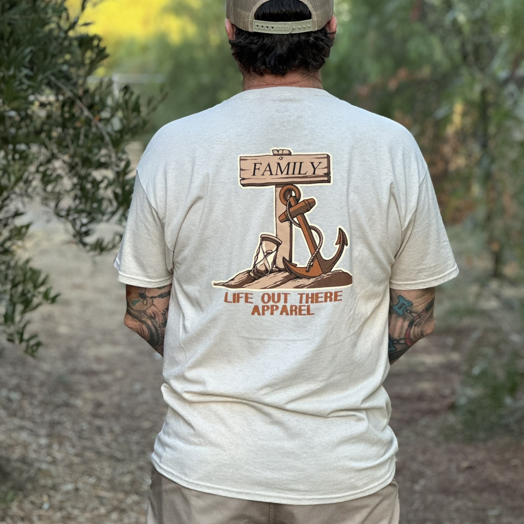 A man dons the Unisex Time Flies T Shirt in the sandstone color, showcasing a design with a wooden sign, rope, and anchor embellished with the words "FAMILY" and "LIFE OUT THERE APPAREL." He stands outside against a backdrop of blurred greenery, embodying the timeless message that time flies when cherished with loved ones.