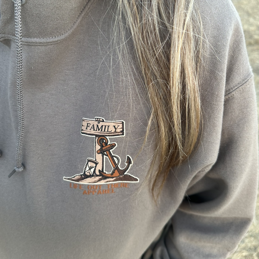 A person wearing the "Unisex Time Flies Hoodie - Safari" by Life Out There Apparel, featuring an anchor design and a wooden sign with the word "Family," along with the phrase "Live on the Hook Apparel" below it. The person's partially visible hair reminds us that time flies when you're anchored in family moments.