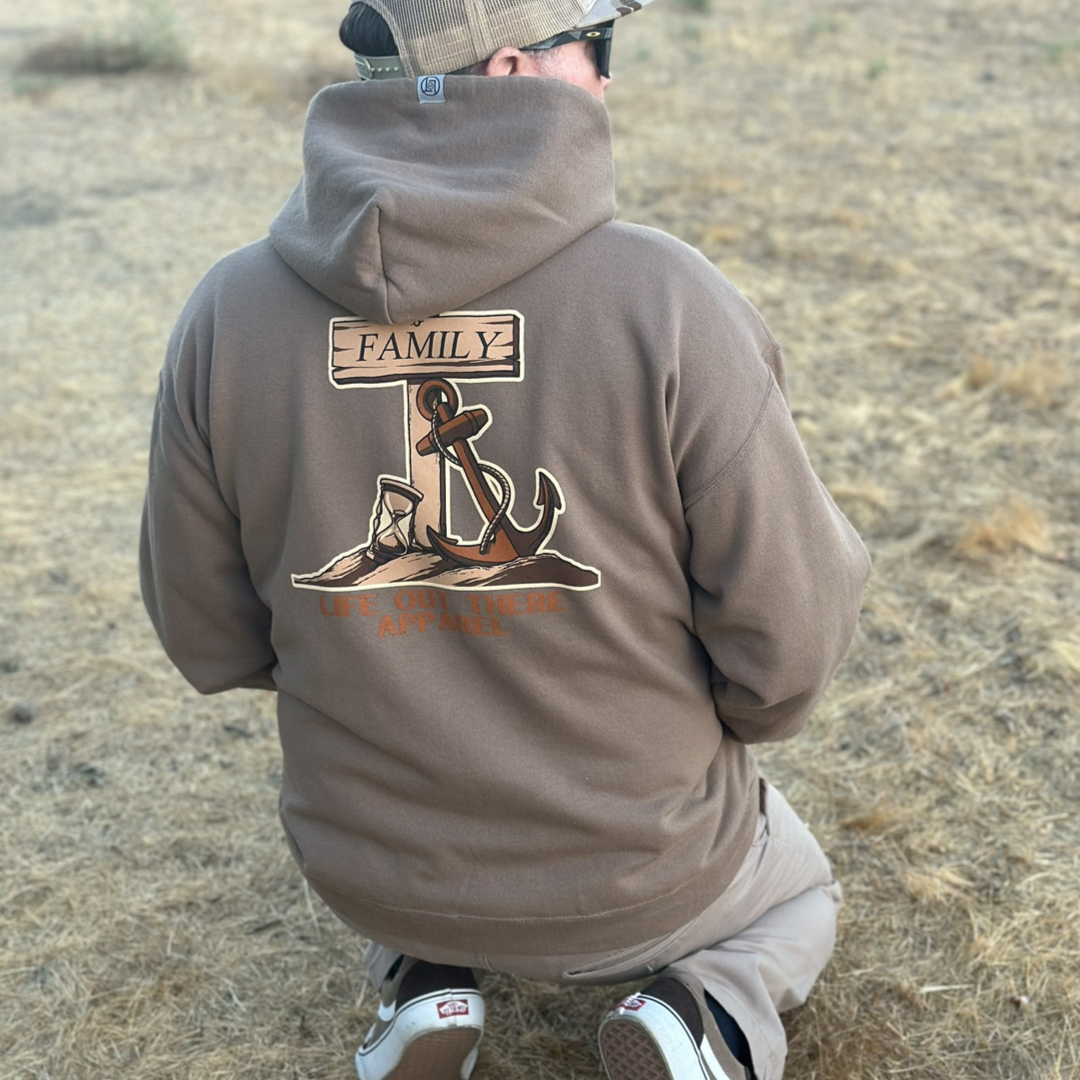 A person kneels on a dry, grassy field, facing away. They are wearing the Unisex Time Flies Hoodie in Safari by Life Out There Apparel. The brown hoodie is decorated with an anchor graphic and features a design with the words "FAMILY" and an hourglass symbolizing that "TIME FLIES." The setting appears to be outdoors on a dry, grassy area.