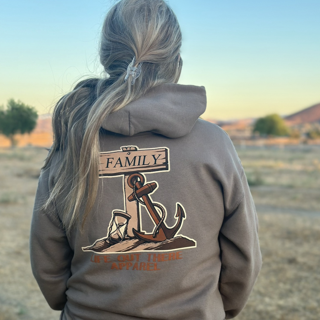 A person with long hair, clad in the Unisex Time Flies Hoodie - Safari by Life Out There Apparel, stands outdoors, facing away. The brown hoodie features an anchor design with the word "FAMILY." As they reflect on how time flies, a dry, grassy landscape extends in the background.