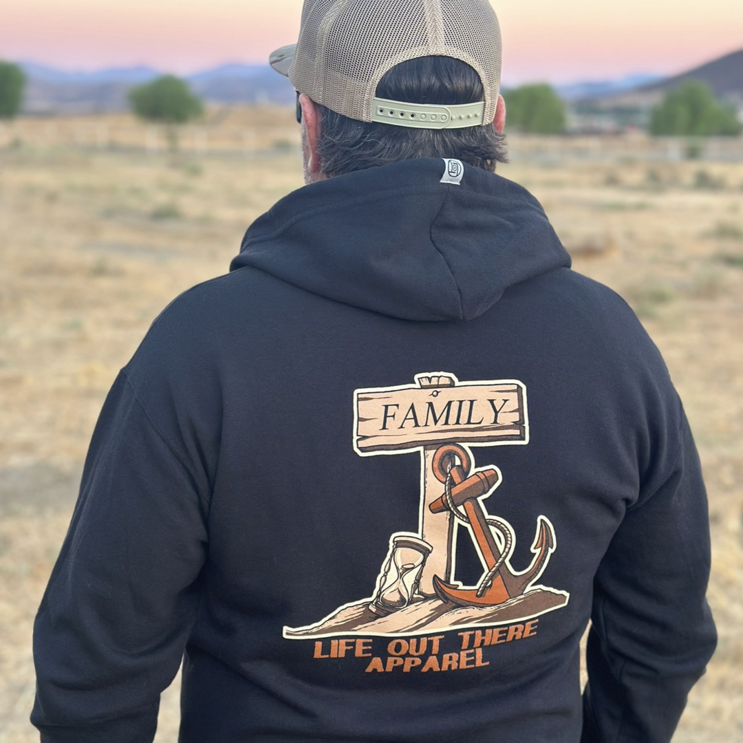 A man in a black Unisex Time Flies "Zip-Up" Hoodie from Life Out There Apparel gazes at the field landscape under the sunset's glow. The hoodie features a design with an anchor, cowboy boots, and a sign that reads "Family," complemented by the phrase "Life Out There Apparel" beneath an hourglass symbolizing Time Flies.