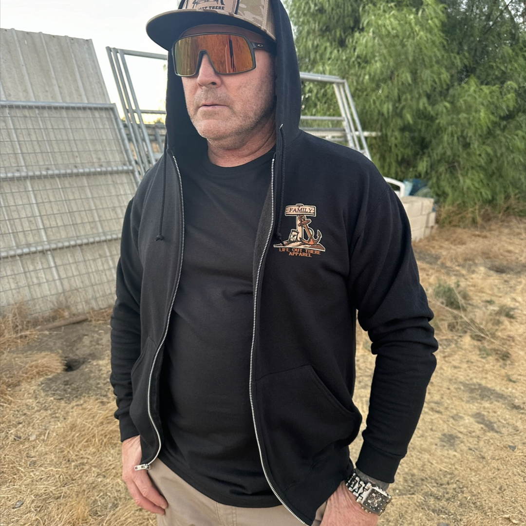 A person wearing the Unisex Time Flies "Zip-Up" Hoodie in black from Life Out There Apparel, along with sunglasses and a cap, stands anchored near a metal fence. They have a serious expression and their hands in their pockets, set against a backdrop of greenery and dry grass.