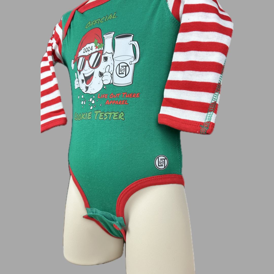Green long sleeve baby shirt with red & white striped sleeves with a cartoon cookie wearing a Santa hat and sunglasses labeled "2024," alongside glass milk cartons. Text reads, "Life Out The Apparel / official cookie tester”.
