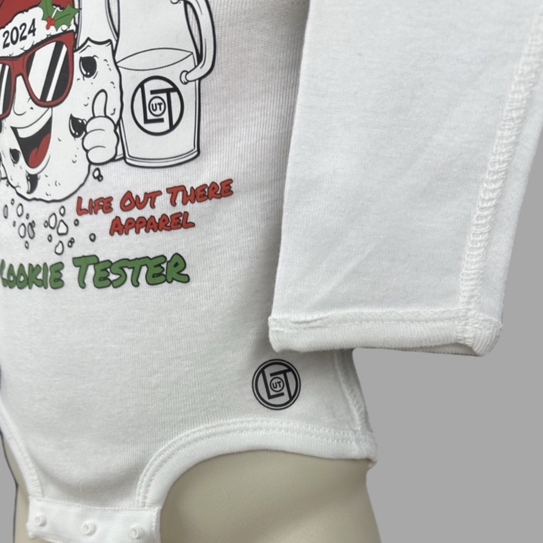 White long sleeve baby shirt with a cartoon cookie wearing a Santa hat and sunglasses labeled "2024," alongside glass milk cartons. Text reads, "Life Out The Apparel / official cookie tester”.