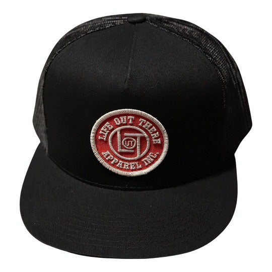 The LOT Snapback "Flat Bill" Trucker Hat in black features a mesh back and is adorned with a red and white circular patch on the front, displaying the text Life Out There Apparel.