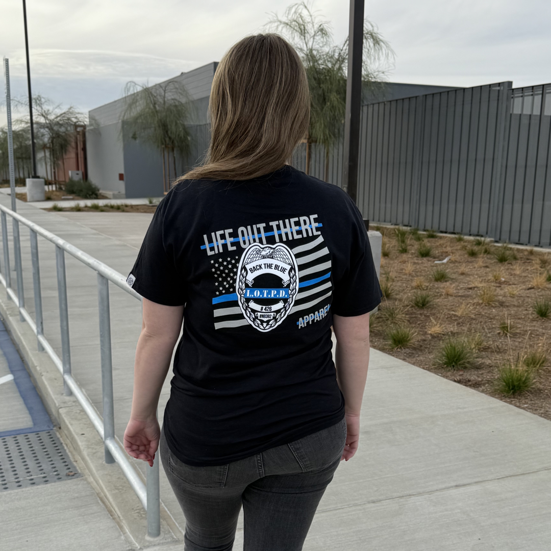 A person in a black Unisex - Back The Blue Tee by Life Out There Apparel stands on a sidewalk. The T-shirt has an American flag design supporting Back the Blue. Modern buildings and sparse trees appear in the cloudy background.