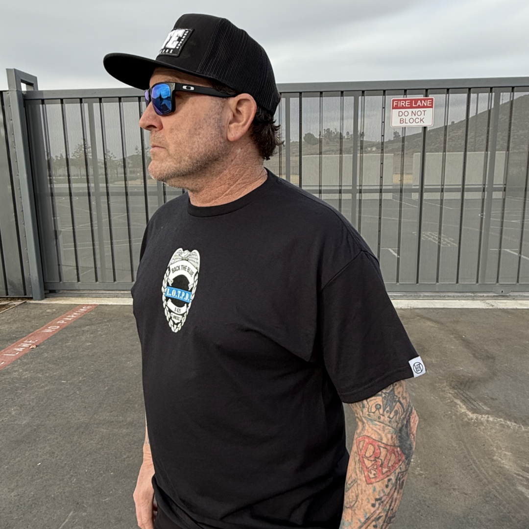 A tattooed man stands outdoors in an overcast sky near a Fire Lane Do Not Block sign, wearing a Life Out There Apparel Unisex Back The Blue Tee in black, a matching cap, and sunglasses by a metal gate, honoring the spirit to support Back the Blue.