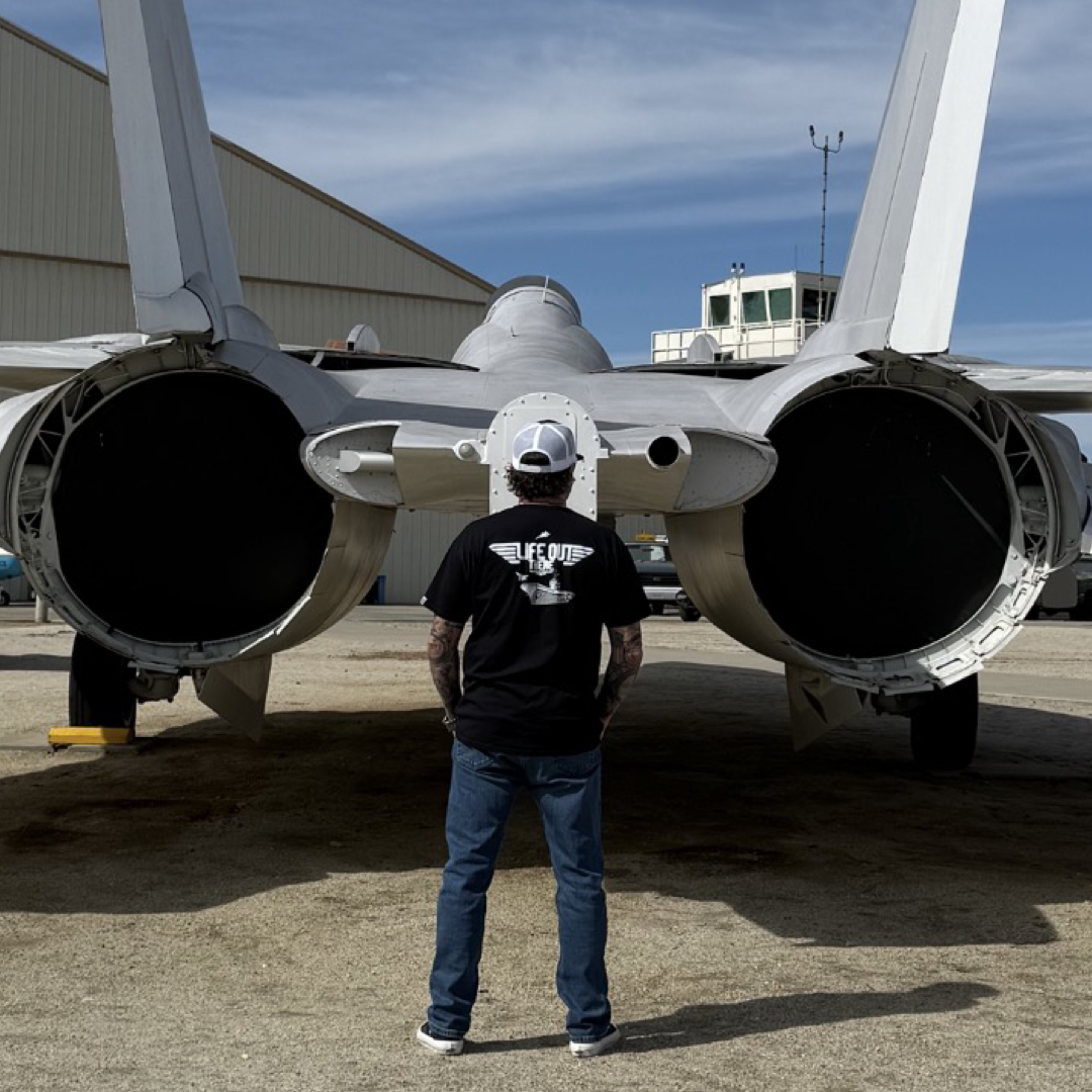 A person wearing the Life Out There Apparels Unisex - The Hard Deck Tee in black and a white cap stands on concrete, facing the rear of a fighter jet. Its large engines and tail fins hint at TOPGUN prowess, while a building looms in the background under a clear blue sky.