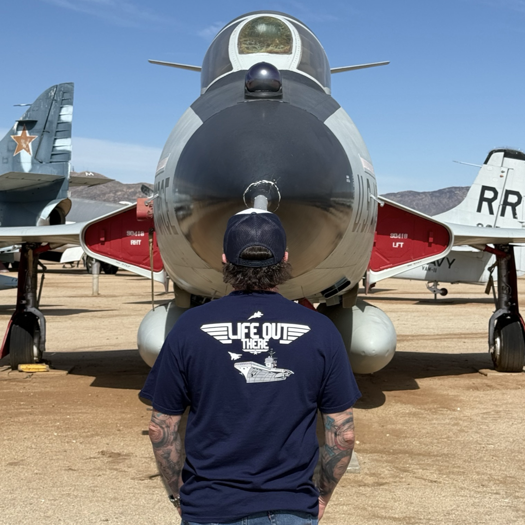 A tattooed individual in a cap, viewed from behind, stands before a vintage jet fighter, evoking a TOPGUN MAVERICK vibe. Theyre wearing Life Out There Apparels Unisex - The Hard Deck Tee in navy, as sunlight highlights the outdoor setting filled with other aircraft.