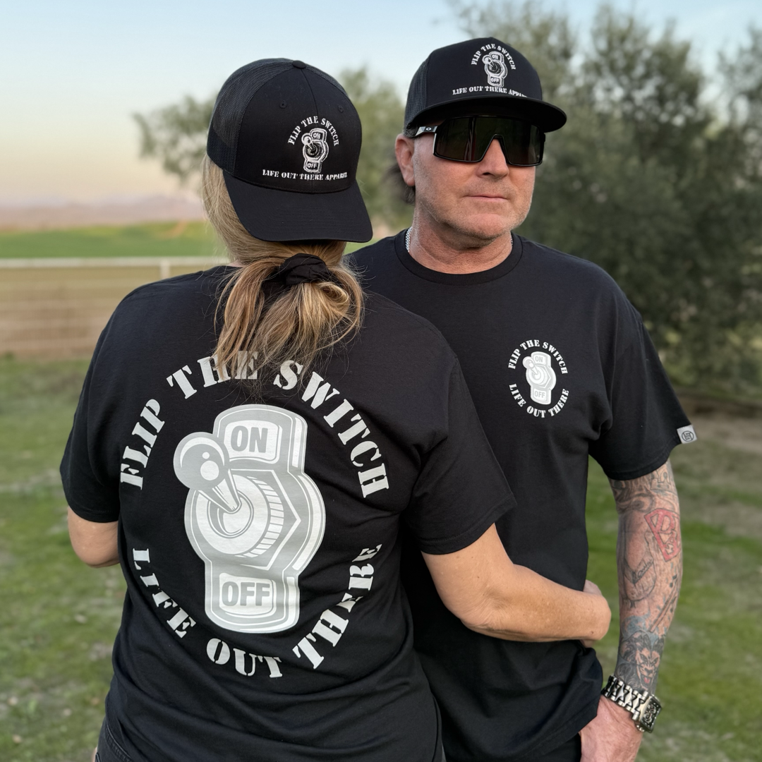 A man and woman stand on grass under trees, both in black Flip The Switch tees by Life Out There Apparel. Their shirts feature an ON OFF switch design and the phrase FLIP THE SWITCH LIVE OUT THERE, paired with stylish sunglasses.