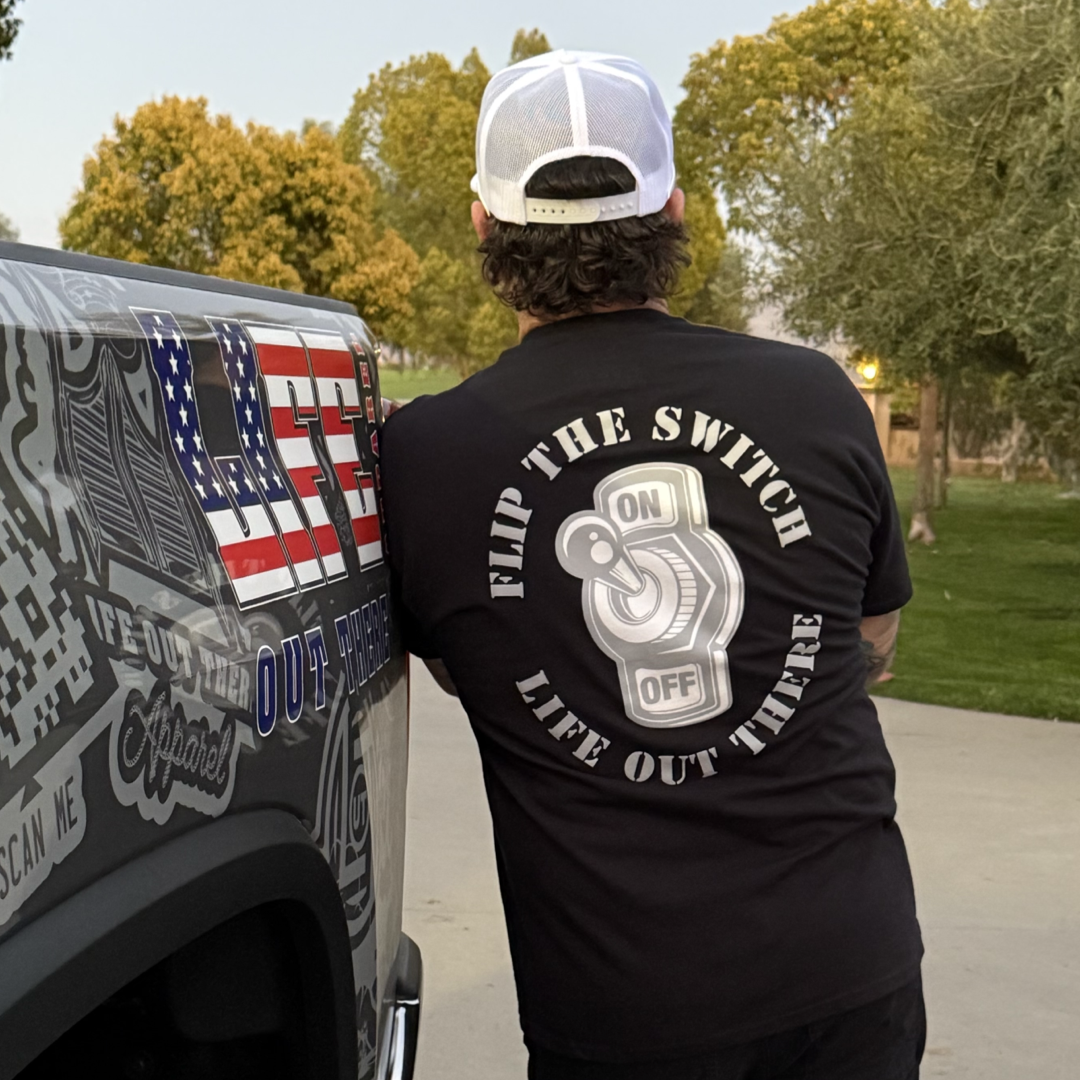 Wearing a Flip The Switch Tee by Life Out There Apparel, someone leans on a vehicle with American flag decals, set against trees and grass.