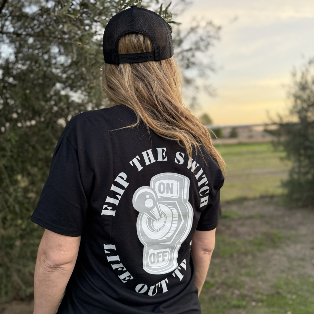 A person outdoors wears the Unisex - Flip The Switch Tee - Black by Life Out There Apparel. The shirt features a bold white switch graphic labeled ON and OFF, surrounded by the phrase FLIP THE SWITCH LIFE OUT TV, set against trees and clear skies.