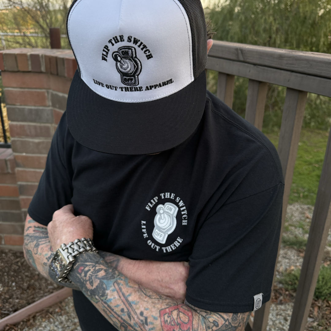 A tattooed person crosses their arms, wearing Life Out There Apparels Unisex Flip The Switch Tee in Black. A black cap complements the look against a wooden fence and brick wall with greenery.