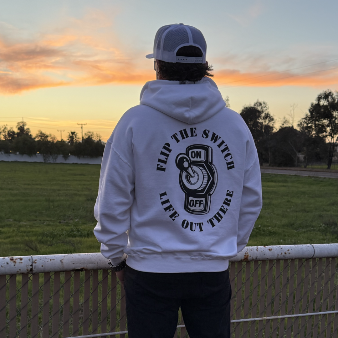 A person wearing the Unisex - Flip The Switch Hoodie in white by Life Out There Apparel, featuring Flip the Switch, Life Out There and an on/off graphic, faces away from the camera. They stand in a grassy field at sunset with a cap on, as trees silhouette against the sky for an anniversary launch celebration.