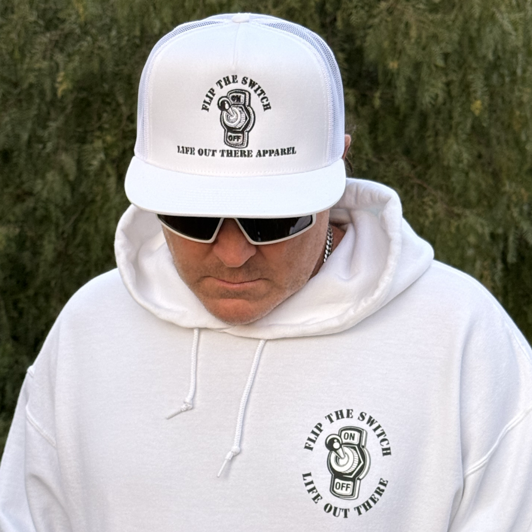 A person in a Unisex Flip The Switch Hoodie by Life Out There Apparel celebrates its anniversary. The white hoodie with a switch design is paired with a matching cap. Sporting sunglasses, the individual gazes downward, contrasted by blurred greenery.