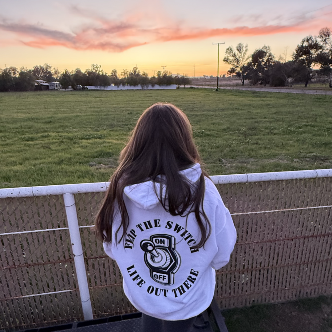 At sunset, a person with long hair stands on a balcony in Life Out There Apparels Unisex Flip The Switch Hoodie in white, featuring a light switch and the phrase FLIP THE SWITCH, gazing over a vast grassy field under vibrant skies and trees.
