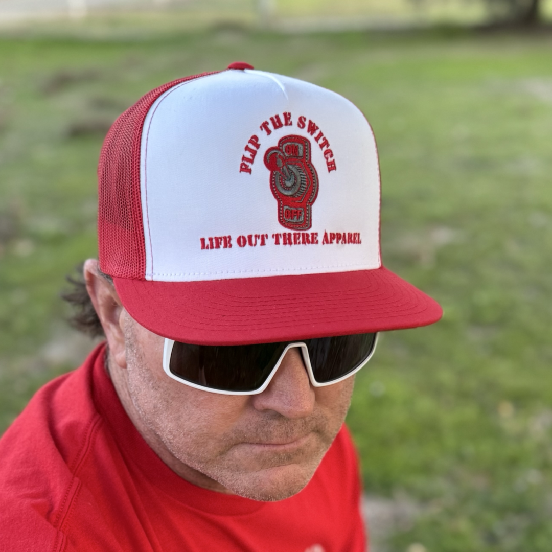 A man outdoors sports a red/white FLIP THE SWITCH flat bill SnapBack trucker hat by Life Out There Apparel, sunglasses, and a red T-shirt, celebrating the brands anniversary with grass in the background.