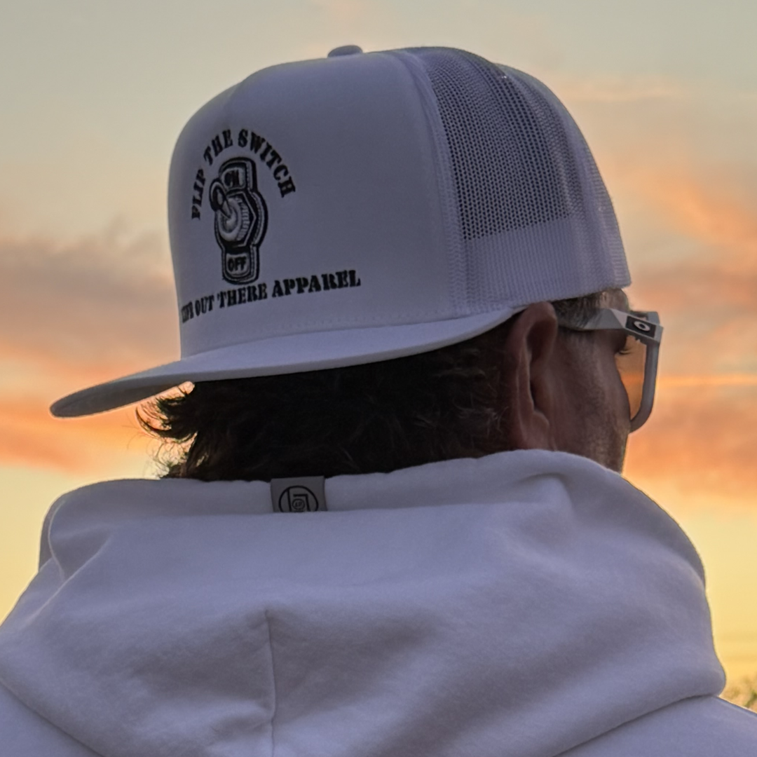 Against a vibrant sunset, a person dons the Life Out There Apparel FLIP THE SWITCH flat bill snapback trucker hat in white. The caps light switch graphic perfectly complements their white hooded jacket.