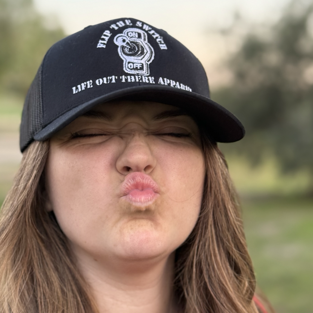 A person with long hair, sporting Life Out There Apparels FLIP THE SWITCH - Curved Bill SnapBack Trucker Hat in black, puckers their lips as if to kiss. The blurred background hints at greenery and open space, capturing the essence of an anniversary launch.