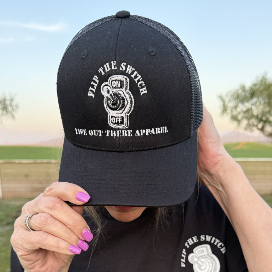 A person wearing Life Out There Apparels FLIP THE SWITCH - Curved Bill SnapBack Trucker Hat in black, featuring OFF ON text, pairs it with a matching T-shirt and purple nail polish against a scenic tree-filled backdrop during the brands 3-year anniversary launch.