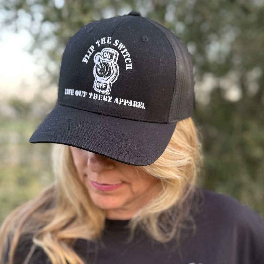 A person with long blonde hair wears Life Out There Apparels FLIP THE SWITCH - Curved Bill SnapBack Trucker Hat in black, featuring the slogans FLIP THE SWITCH and ON/OFF, for their anniversary launch. The individual gazes downward against a backdrop of blurred greenery.