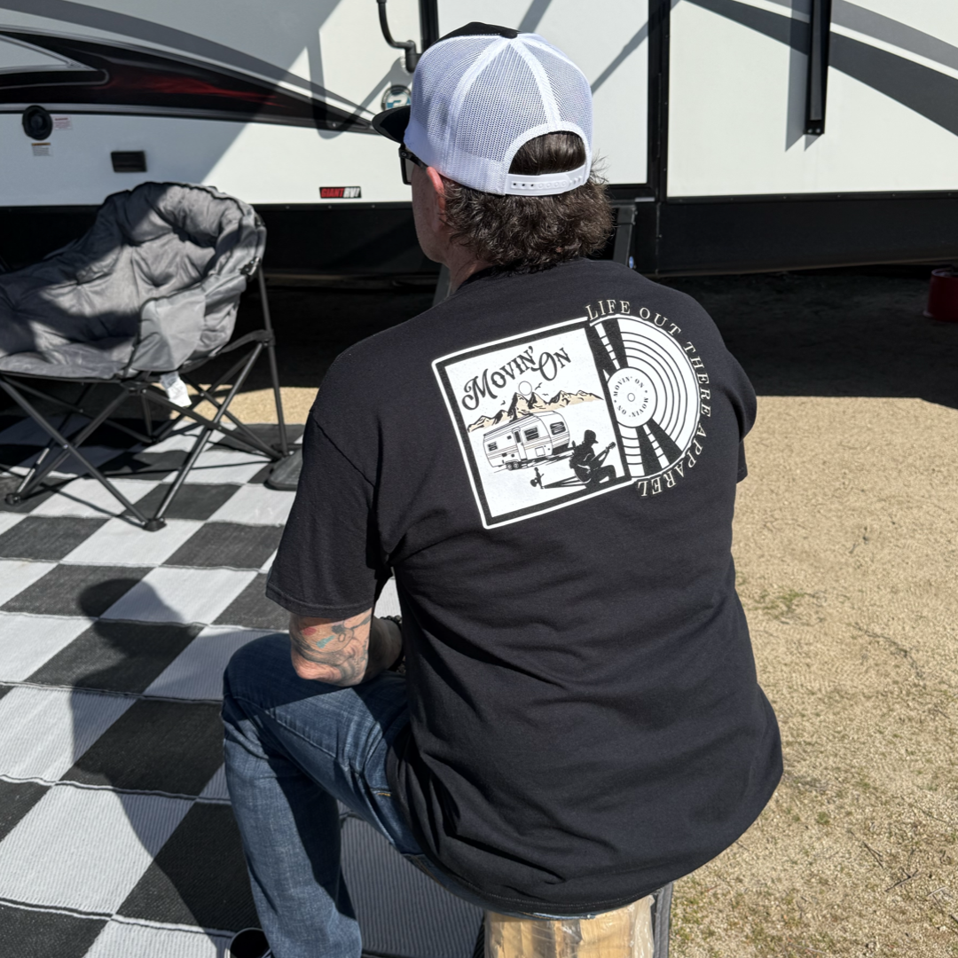 A young man in a Life Out There Apparel Unisex - Movin On Tee - Black and a white cap sits on a wooden bench outdoors, with a folding chair and part of an RV nearby on a checkered mat—a country album cover moment.