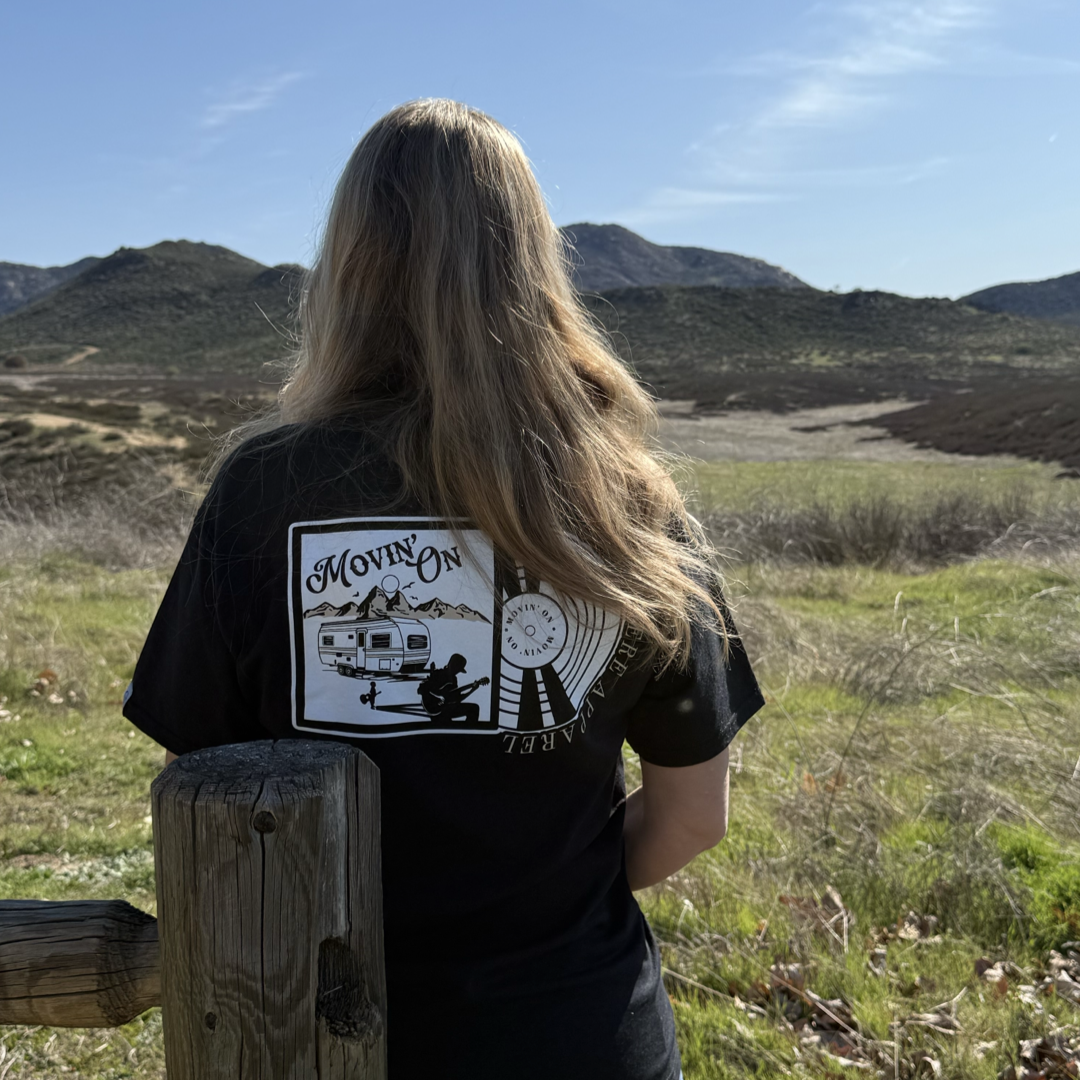 A person with long blonde hair, wearing the Life Out There Apparel Unisex Movin On Tee in black with a camper, sun, and MOVIN ON graphic on the back, stands before green hills and mountains by a wooden fence.