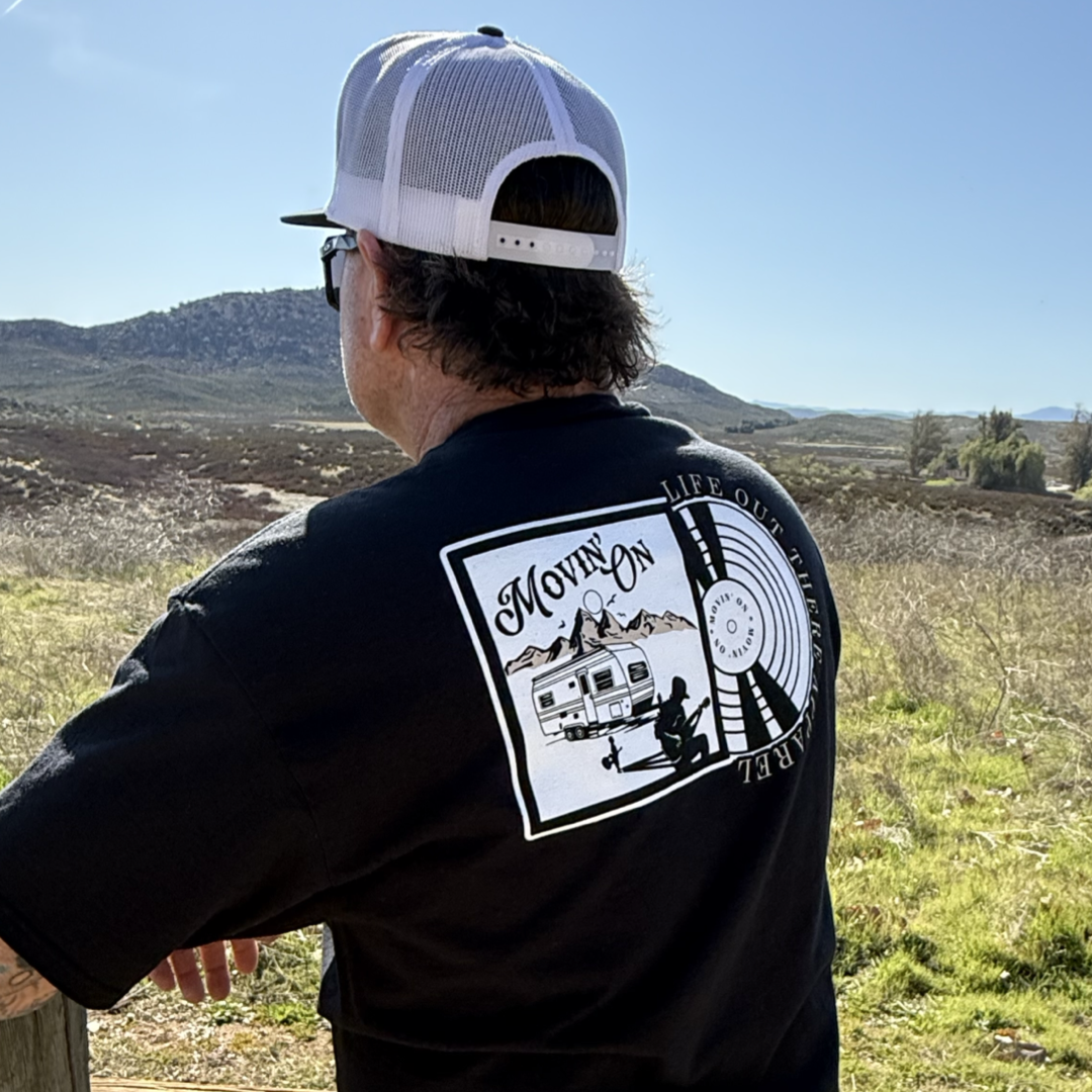 Wearing the Unisex - Movin On Tee - Black by Life Out There Apparel, and a white mesh cap, someone stands outdoors gazing at scenery. The tee features “MOVIN’ ON,” a camper, mountains, and "Life Out There Apparel" — perfect for album covers.
