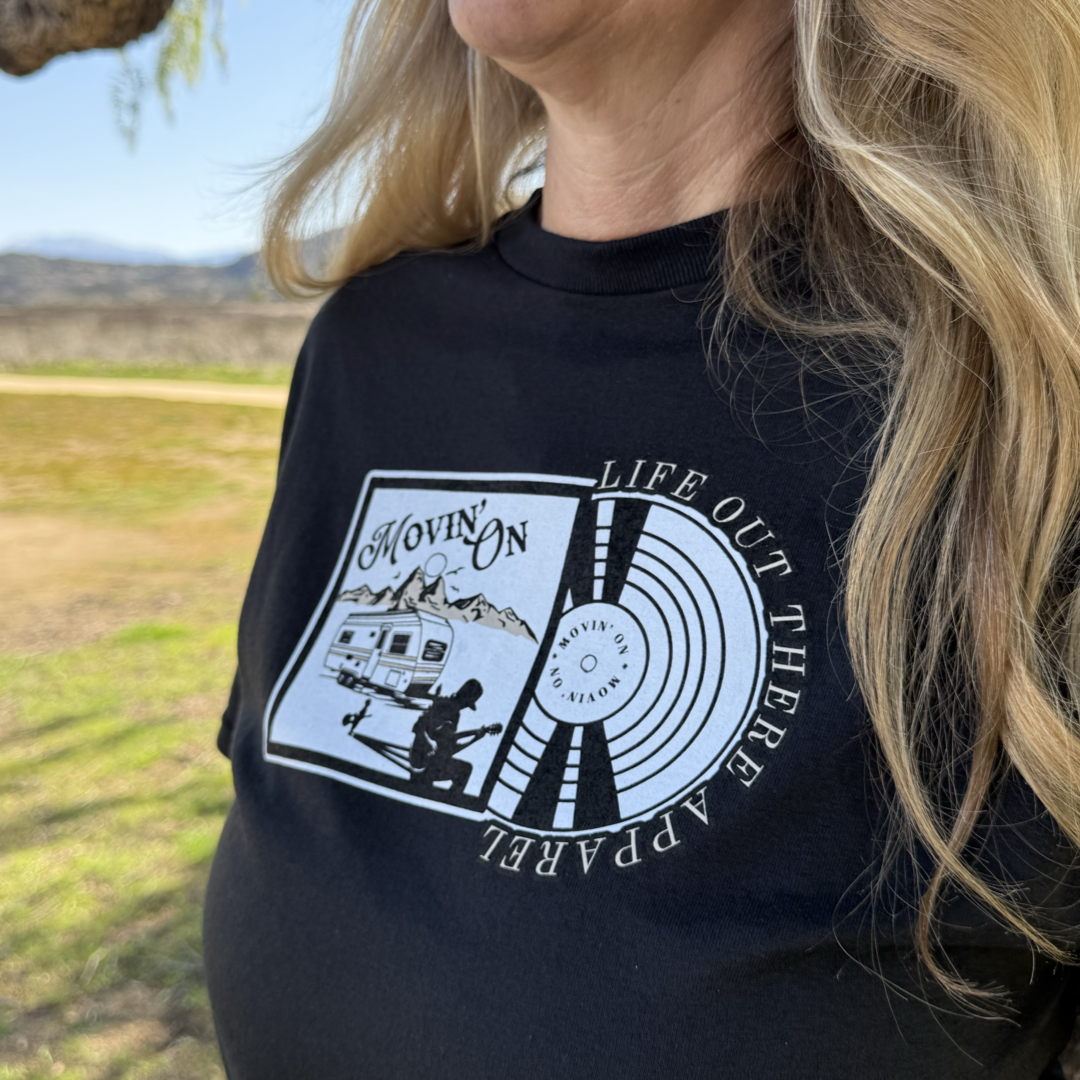 A young woman wears the Unisex Movin On Tee in black by Life Out There Apparel, featuring a camper, mountains, guitar player, and vinyl record design with “Movin On” text—an album cover style evoking country music and adventure.