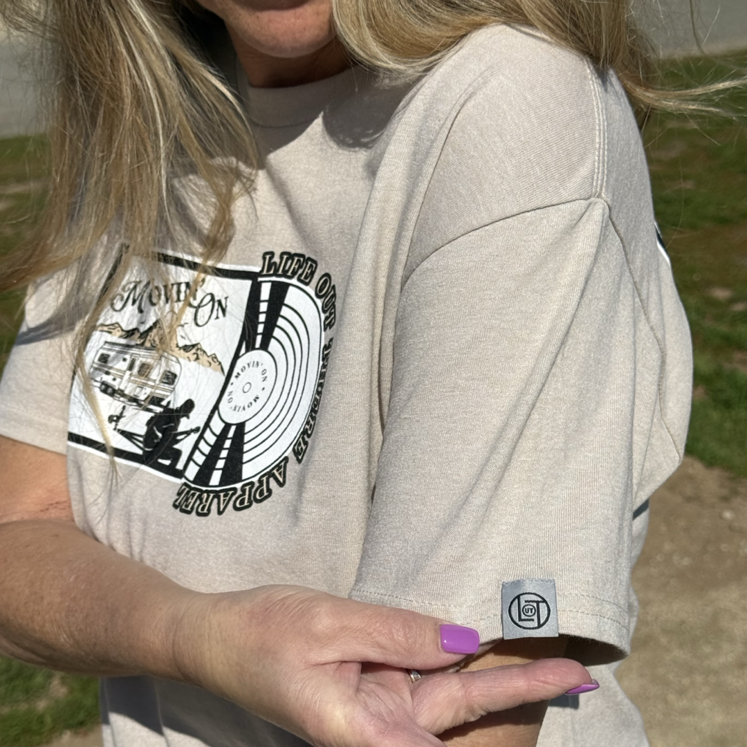 A woman with long blonde hair points to the logo tag on her beige Unisex Movin On Tee - Sandstone by Life Out There Apparel, featuring a graphic trailer and vinyl record design with MOVIN ON and Jeremiah Watson.