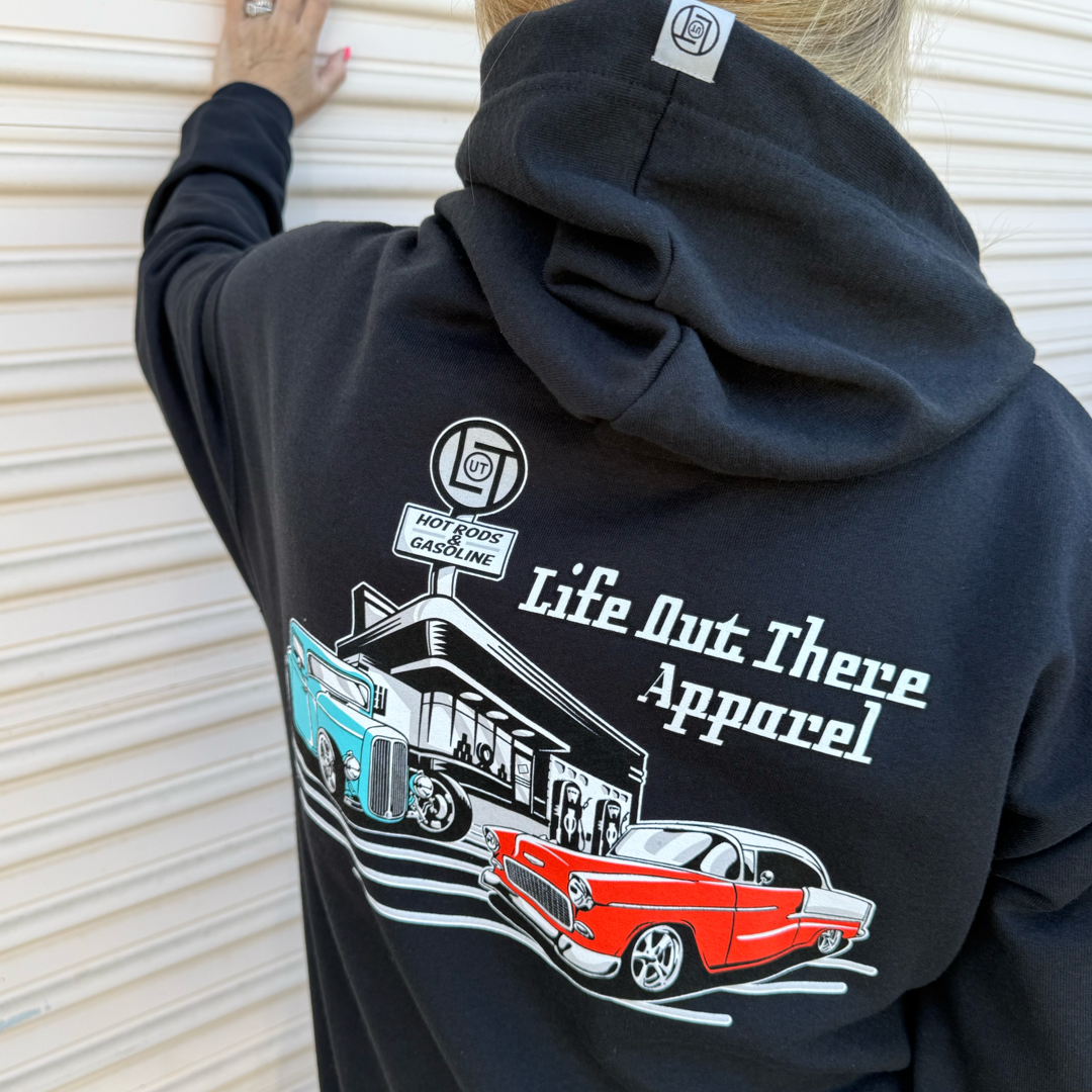 A person wears the Unisex Cruzin Zip-Up Hoodie in Black by Life Out There Apparel, featuring a back graphic of a vintage gas station, blue hot rod, and red Chevy Bel Air—ideal Fathers Day gift.