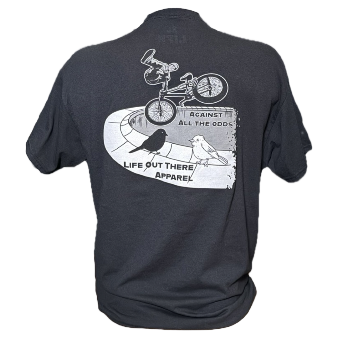 The Unisex Against All The Odds / The Ride Tee in black by Life Out There Apparel features BMX legend John Buultjens performing a pool trick, bold text AGAINST ALL THE ODDS, and two bird illustrations.