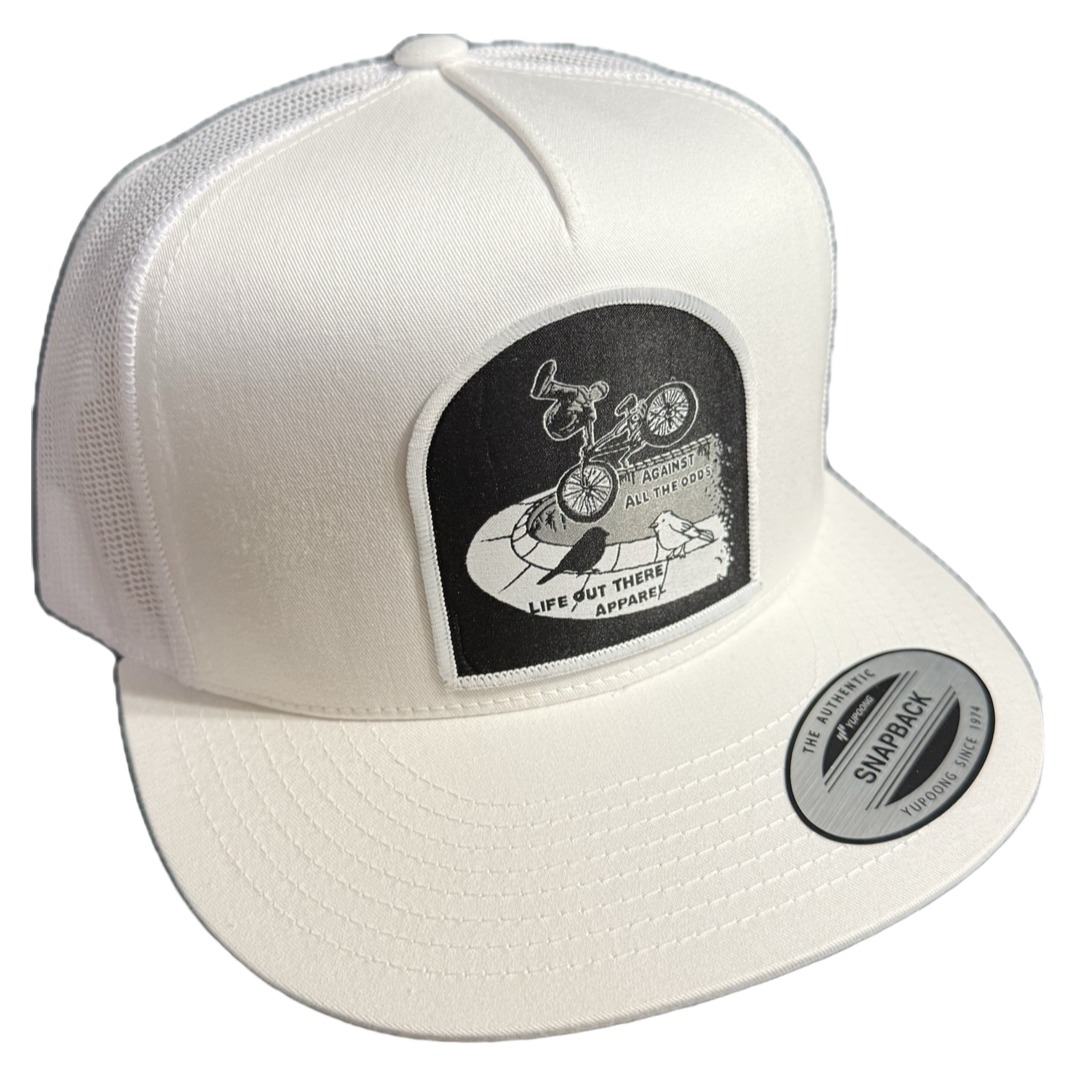 The Against All The Odds / The Ride Flat Bill SnapBack Trucker Hat in white by Life Out There Apparel features a mesh back, a black and white bicycle patch, and unique illustrations—ideal for fans of THE RIDE or BMX pro John Buultjens.