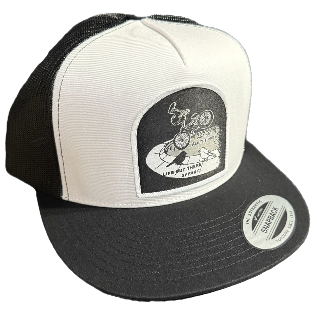 The Against All The Odds / The Ride SnapBack Trucker Hat by Life Out There Apparel features a white/black design, mesh back, flat bill, and a circular patch with BMX legend John Buultjens, cyclist art, and inspiring text.