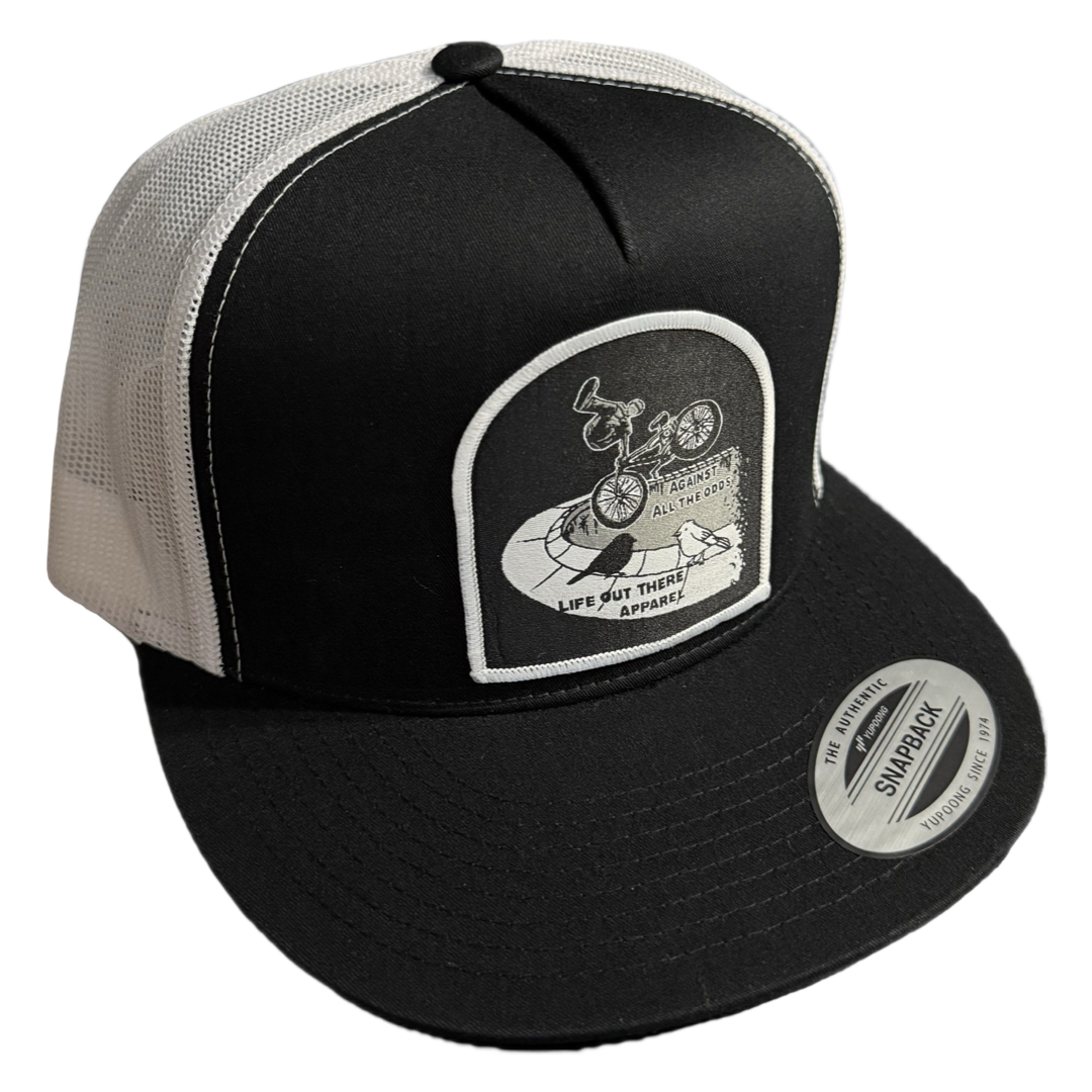 The Against All The Odds / The Ride Flat Bill SnapBack Trucker Hat in black and white by Life Out There Apparel features a mesh back, BMX rider patch, Life Out There Apparel text, and an The Authentic Snapback sticker on the brim.