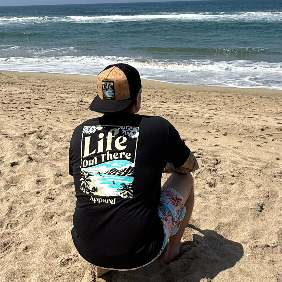 Wearing the Unisex Aloha Tee in black by Life Out There Apparel, a person in a cap sits on the sandy North Shore of Oahu, gazing at waves and a blue sky, soaking up Hawaiian vibes.